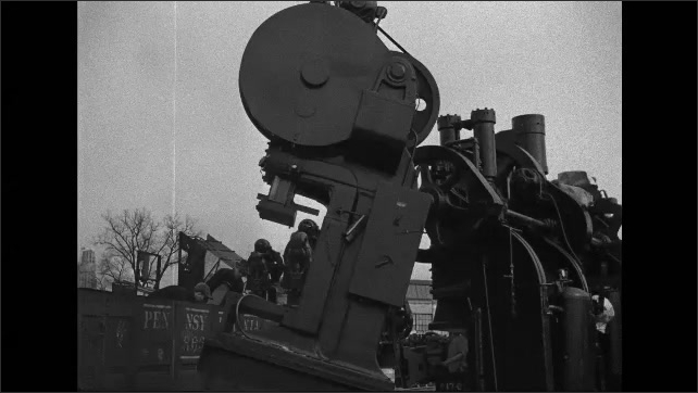 1940s: Crane lowering punch press. Men in rail car hoisting object over side of car onto ground. Punch presses outdoors.