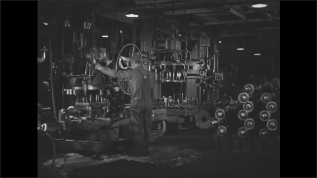 1930s: A piece of metal is adjusted and multiple drills continue to drill holes. Worker watches a drill jig operate. Two workers stack an axle from one station and move another to a new station.