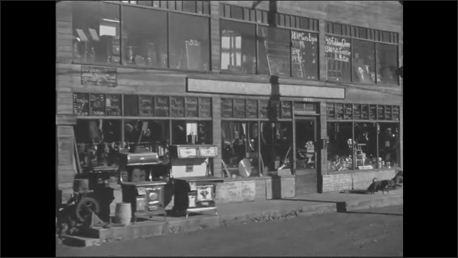 1930s: Car driving past a used item store and lot. Slate. Junk cars stand in a field with buildings.