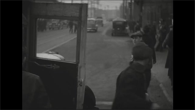 1930s: Men step off bus and walk along crowded sidewalk in city. Men carry lunch bags and enter factory yard.