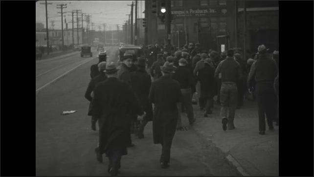 1930s: Men walk along crowded sidewalk in city. Men carry lunch bags and enter factory yard.