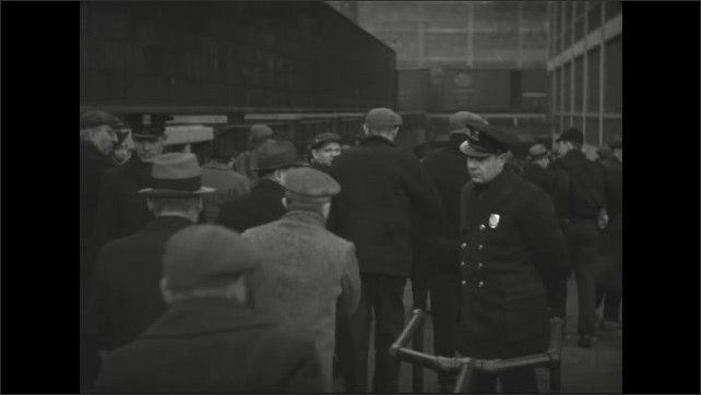 1930s: Men carry sack lunches past security guards and enter factory yard.