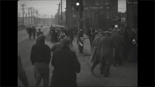 1930s: Men carry sack lunches past security guards and enter factory yard.