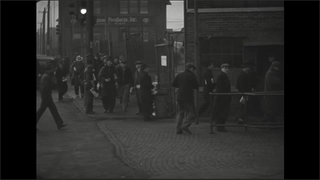 1930s: Men step off bus and walk along crowded sidewalk in city. Men carry lunch bags and enter factory yard. Hand holds production slate near factory yard entrance.