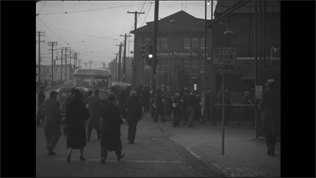 1930s: Men and women step off bus and walk along crowded sidewalk in city. Workers carry lunch bags and enter fenced factory yard.