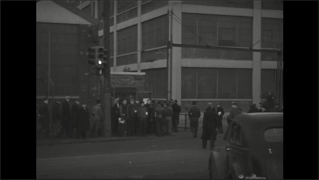 1930s: Men and women cross busy street and walk along crowded sidewalk in city. Workers carry lunch bags and enter fenced factory yard.
