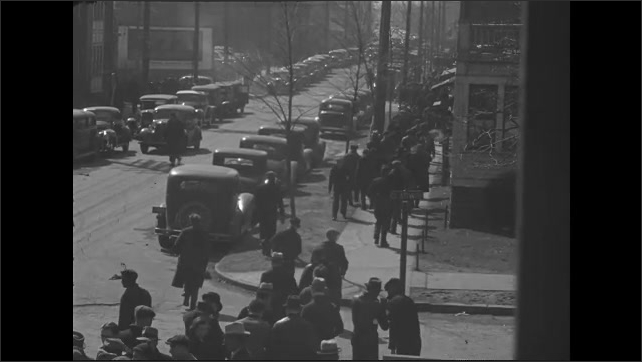 1930s: People walk through crowded city streets toward factory. Guard opens doors to factory and workers walk in.