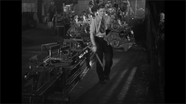 1930s: Worker with hoist moves an engine onto a conveyor belt. Worker moves the hoist. Worker attaches a clamp to an engine, taps the bottom and places it on a conveyor belt. Slate. Stored engines.