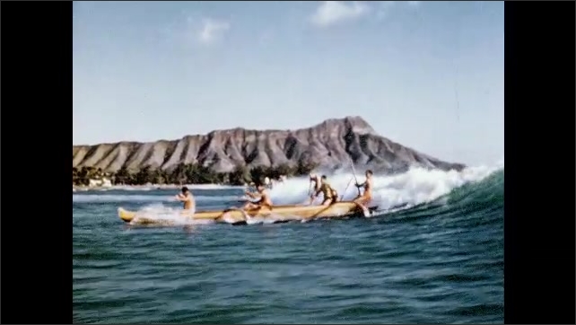 1950s: Outrigger canoe and surfers paddles towards shore. Wave crests and surfers stand on board.