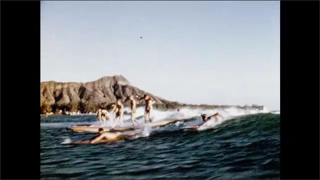 1950s: Single surfers ride board. Woman does shoulder mount on surfer on surfboard. Couple stand on surfboard and surf. Man falls off surfboard into water.