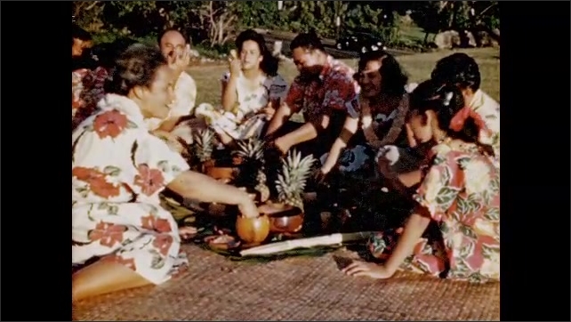 1950s: Group of seated people eat food on a blanket, including poi. Busy streets of Honolulu with traffic and buses. 