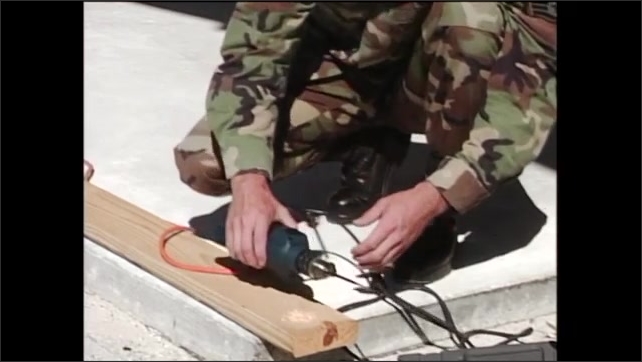 2000s: After drilling hole in wood plank, man in army fatigues lays drill down then begins loosening drill head.