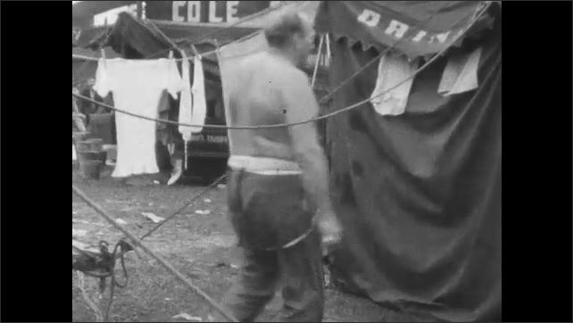 1940s: Black man seated by circus tent gives treat to dog; burly, shirtless man walks into tent, dons shirt.