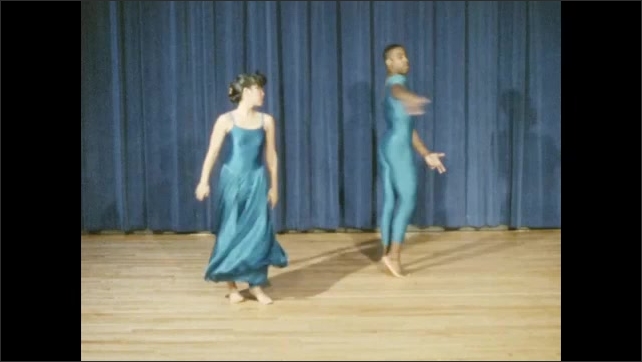 1980s: Teen boy in blazer stands and speaks from an auditorium where actors are rehearsing on stage. Two dancers in blue come forth and put on a beautiful modern dance routine.