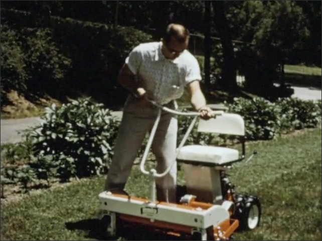 1960s: A riding lawnmower on a lawn. Side view of the chassis. Man grabs mower handles and wiggles it back and forth, sits in the seat. The engine. Man pulls rear lever.