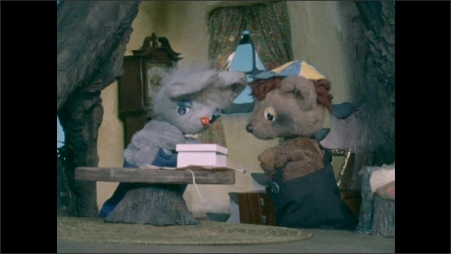 1970s: Puppet theater, treehouse living room, rabbit stands over box, bear talks, nods head. Rabbit lifts lid, bear looks inside box, rabbit reads paper. Shirtless human man pops up, talks.