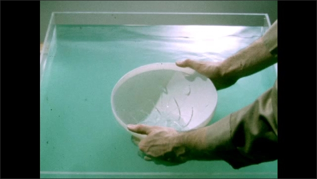 1950s: Hand holds chalk slate. Hands push bowl with holes into water. Water fills bowl through holes in surface. Colored fluid rests in fluid pressure device on table.
