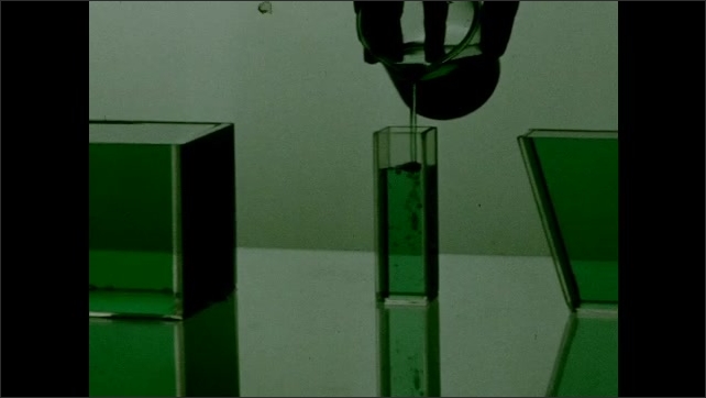 1950s: Colored fluid rests in glass rectangle on table. Hand pours colored fluid into glass cylinder on table. Colored fluid fills glass shapes on light table. 
