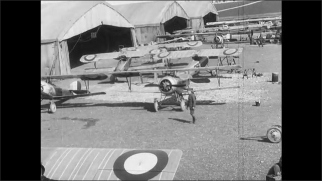 1910s France: Airplanes sit at hangars.  Men stand near plane.  Man moves propeller and speaks.