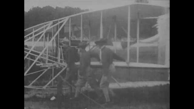 1900s: Several people carry Wright Brothers airplane across field, and place it on tracks. Intertitle card. Orville Wright sits in pilot's seat.