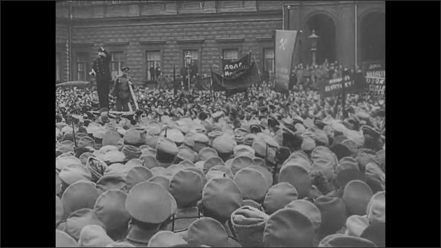 1910s: Large crowd of people. Man on platform addresses crowd. Officer on platform addresses crowd. Light from balcony window at night.