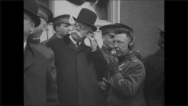 1910s: Men and generals look up together, watching something. Man speaks into microphone, man next to him wears headphone. Generals and officers.