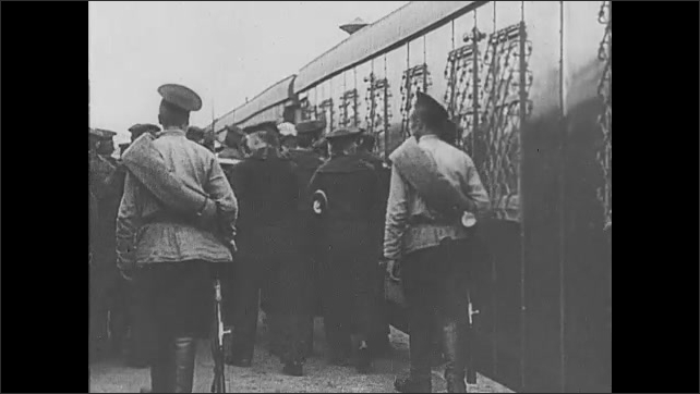 1910s Russia: Man in long coat stands in middle of circle of marching men. Men behind bars with guard. Men with rifles march prisoners on to trains.