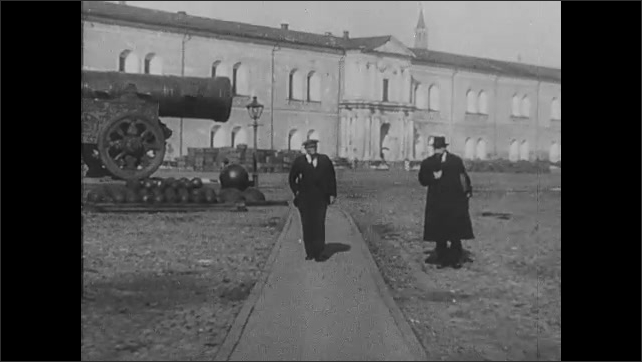 1910s Russia: Lenin stands outside speaking collegiately with another man. Lenin stands in front of cannon and building. The two men walk and talk. 