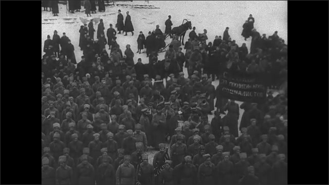 1910s USSR: Soldiers in long coats on horseback ride through large crowd. Lenin speaks loudly in Russian to a crowd of people in head kerchiefs. 