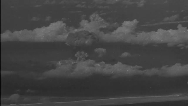 1940s Bikini Atoll: Explosion and shockwave spread across island. Mushroom cloud rises into air.