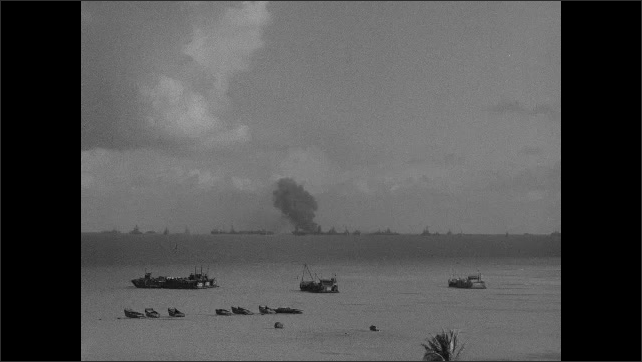1940s: Boats sit stationary in ocean with fleet of Navy ships on horizon as fallout and debris from nuclear explosion settles in distance. Smoke rises rapidly from Navy ship.
