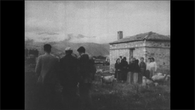 1950s Greece: Shepherds drive sheep through village.  Groups of people talk.  Women make yarn from wool.