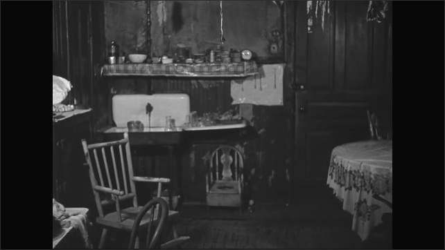 1930s: A well worn kitchen with old fashioned stove and sink. Pots and percolator on the stove. Mismatched chairs and wires from the ceiling.