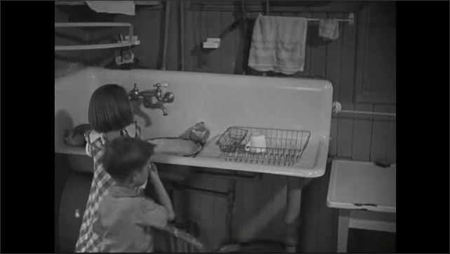 1930s: Girl moves some dishes. She runs water into the pan. She washes a mug. Boy grabs a towel. He starts to dry cutlery. Girl wipes off table.