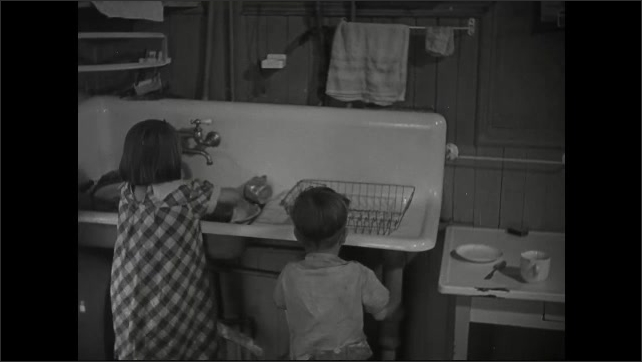 1930s: Boys dries dishes and places them on a table. Girl washes items and places them in draining rack. Toddler naps on a bed.