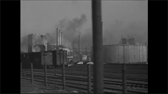 1930s: Car drives along a street, passing a locomotive blowing its whistle. Factory and box cars. Steam rises from a factory.