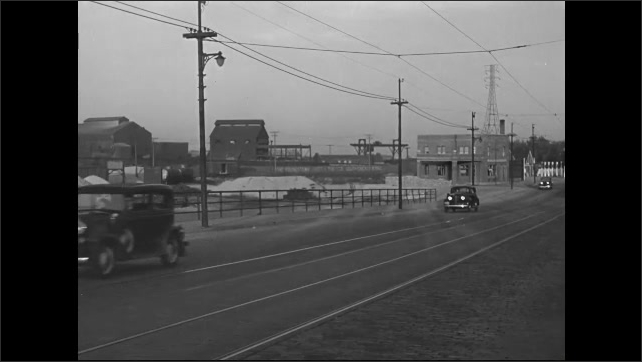 1930s: Cars drive along a road with trolley tracks going down the center. Factory with piles of material in front.