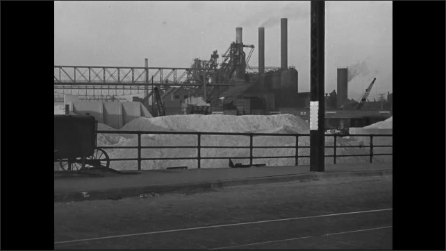 1930s: Cars drive along a road with trolley tracks going down the center. Factory with piles of material in front. Loading dock next to a waterway.