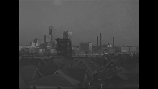 1930s: Row of houses with fences and small yards. City view of house rooflines and chimneys with river in middle. City skyline and factory smokestacks.