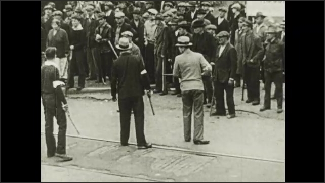 1910s: Men and women walk along sidewalk near factory. Strike busters carry guns and batons down street. Strike busters threaten crowd of workers on street.