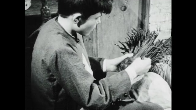 1940s: Boy takes a strip of wood and wraps the whittled strips of a wood broom. Woman places a log on a fire. Boy wraps the broom head. Boy gives the broom to the woman. The woman sweeps.