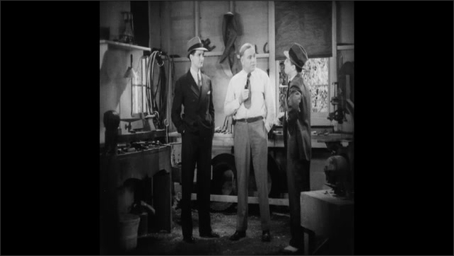1930s: Men sitting at office desk, talking. Men standing in workshop, talking. Man holding hand in in pocket, sucking on tobacco pipe. 