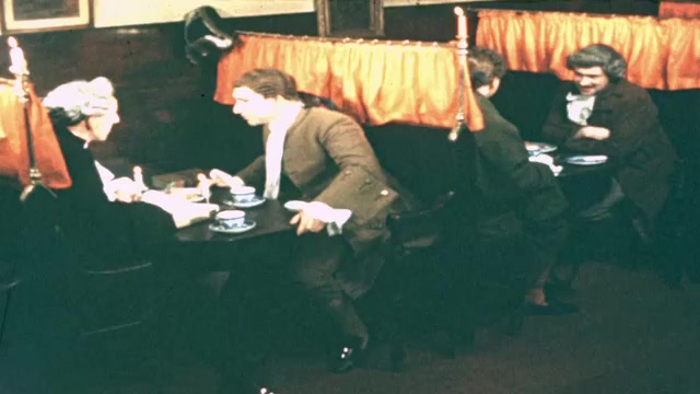 1730s: Restaurant.  Men sit in booths and talk.  Man sips tea.  Narrow street.  Man holds chalkboard shooting slate.  Man looks in store window.