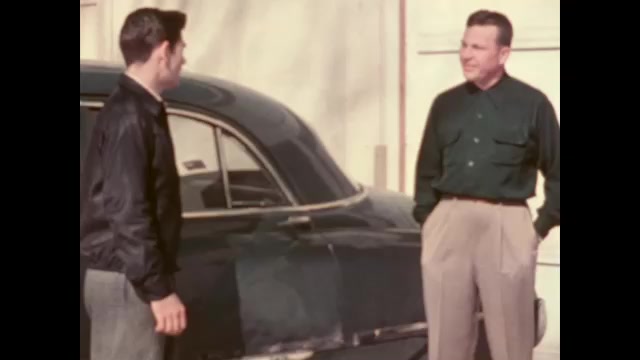 1950s: A man and a teenage boy stand next to a green sedan and talk. The man stoops down and stands back up, speaks to the boy.