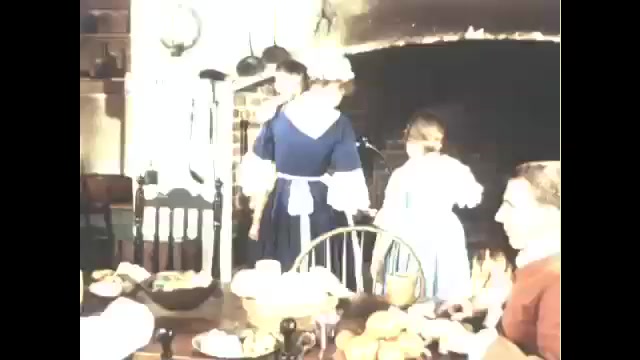 1760s: Family in period costume in kitchen, woman serves dish, sits at table, family eats at table. 
