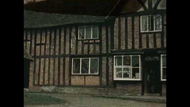 1500s: Man and woman in period costume exit house, people in costume walk down road. Low angle, man and woman exit house. 