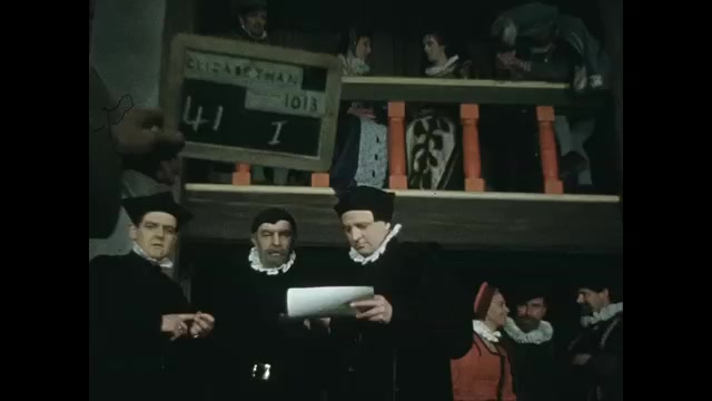 1500s: Hand with clapboard. People in period costume in hall, man writing. Men talking, man hands scroll to man in balcony. 
