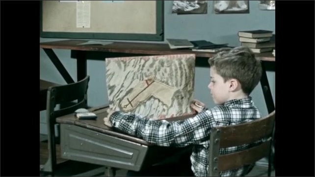 1950s: Boy looks at booklet at desk, pan to boy drawing, holds up drawing. Kids in classroom stand up from desks, exit. 
