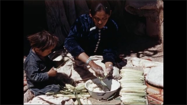 1940s: Woman rolls maize and flour with stone. Woman stuffs maize leaves with dough and lines them up as child sits next to her.
