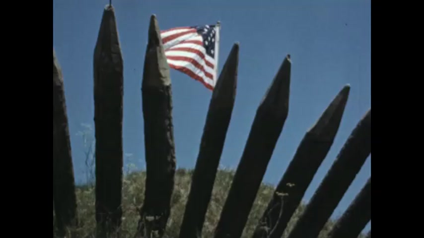 1770s: American flag on pole waves behind wooden spikes. Film slate on ground. British flag on pole behind wooden spikes. Person waves white flag. British flag falls down.